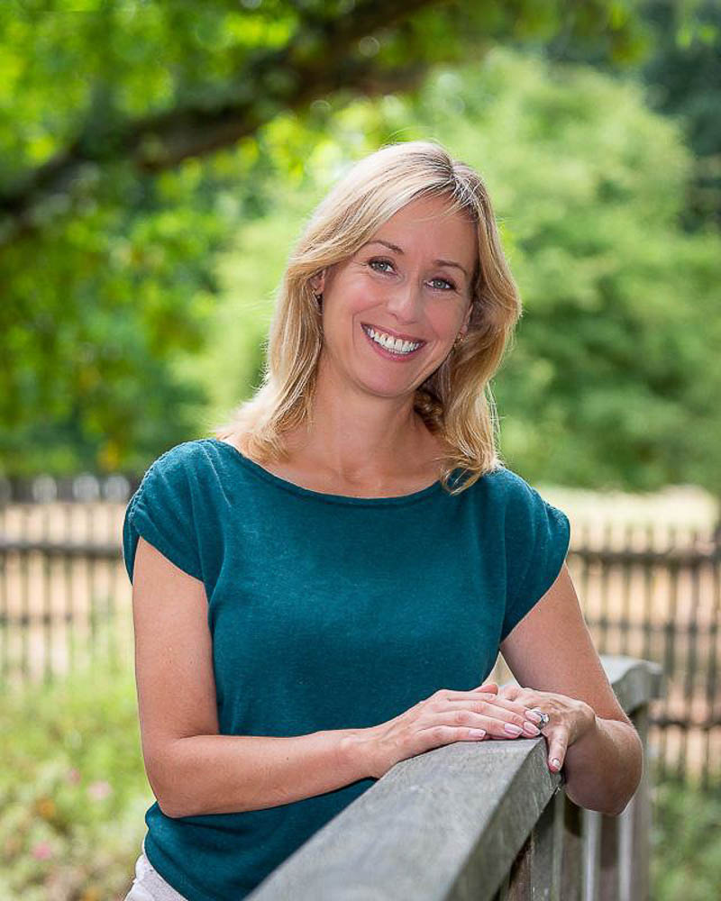 Smiling woman standing outdoors with greenery in the background.